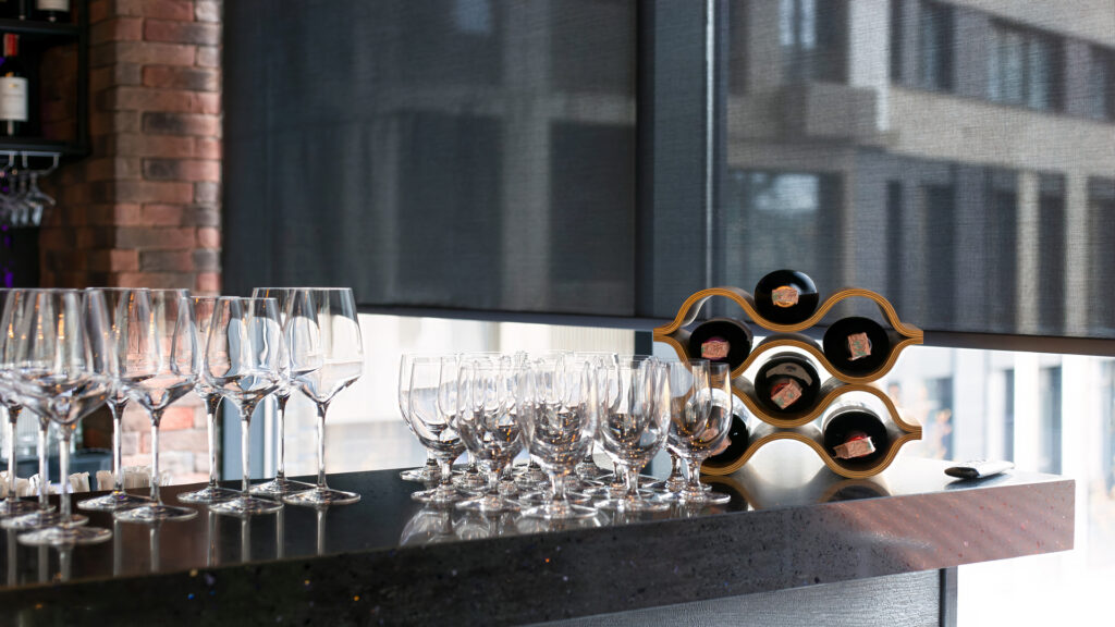 Roller blinds on the window in the restaurant interior. Motorized solar shades, black screen fabric. A control panel for blinds is on the bar counter of the restaurant, glasses and bottles of wine.